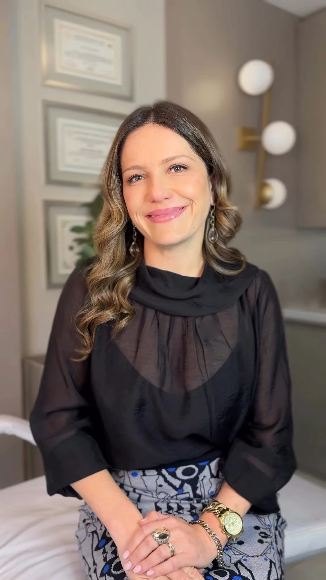 Dra. Leandra Bittencourt sentada no consultório, de blusa preta, sorrindo; na parede atrás dela, diplomas emoldurados e uma arandela de globos.